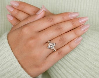 May include: A gold engagement ring featuring a large, square-cut diamond. The ring is adorned with smaller accent diamonds. The ring is on a hand with manicured nails. A symbol of commitment and love.