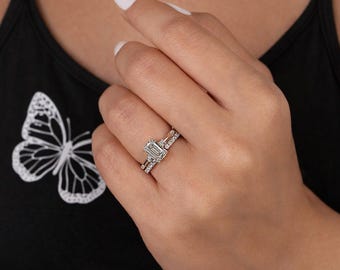 May include: A hand wearing a set of two diamond rings. The engagement ring features an emerald-cut center stone with a halo of smaller diamonds. The wedding band is a delicate row of diamonds. The rings are set in a silver metal.