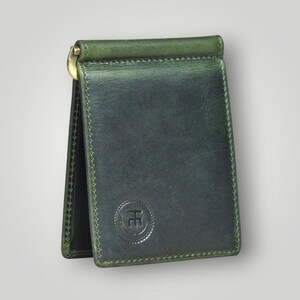 May include: A dark green leather money clip wallet with a gold-colored clasp. The wallet has a stitched border and a circular embossed logo. The leather has a slightly distressed appearance.