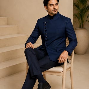 May include: A navy blue bandhgala jacket featuring a high mandarin collar and an asymmetrical front closure with a row of dark buttons. The jacket is paired with matching navy blue trousers, a dark turtleneck sweater, and black leather dress shoes.