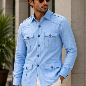 Pode incluir: Uma camisa de manga comprida azul-clara com botões, quatro bolsos frontais com abas, costuras decorativas nas bordas dos bolsos e nos ombros, e botões castanhos escuros, combinada com calças brancas simples.