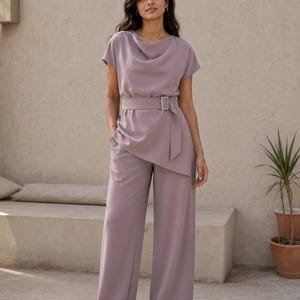 May include: A mauve jumpsuit featuring a cowl neckline, short sleeves, and wide-leg trousers. The garment includes a matching fabric belt with a rectangular silver buckle at the waist, creating an asymmetrical layered effect over the hips.