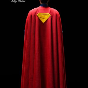 May include: A red cape with a yellow Superman logo, viewed from the back. The cape is draped over a figure with dark hair, set against a black background. The Superman logo is a yellow 'S' inside a yellow diamond.