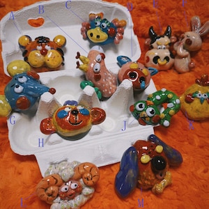 Puede incluir: Una colección de figuras de animales de cerámica hechas a mano, coloridas, que incluyen un tigre, un cangrejo, una vaca, un conejo y un perro, exhibidas en una huevera. Las figuras están pintadas con colores brillantes y detalles caprichosos.