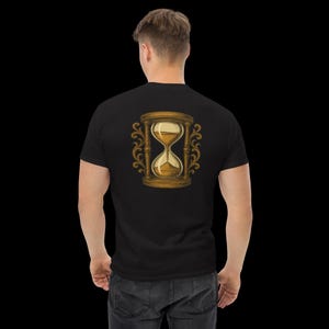 Op de afbeelding: Zwart T-shirt met een gouden en bruine zandloper op de rug. De zandloper is omlijst met decoratieve krullen. Het T-shirt is gemaakt van een zacht materiaal. Het ontwerp is gecentreerd.