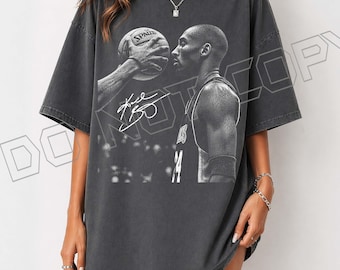 May include: Dark grey oversized t-shirt with a black and white image of a basketball player holding a basketball. The image includes the player's signature and jersey number. The shirt has short sleeves and a relaxed fit.