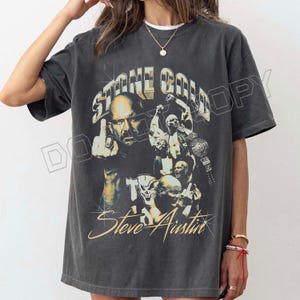 May include: Dark gray t-shirt featuring a graphic of Stone Cold Steve Austin. The design includes images of Austin, the text "STONE COLD" and "Steve Austin", and a championship belt. The shirt has a vintage, distressed look.