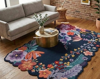 May include: A navy blue, floral-patterned area rug with a scalloped edge. The rug features large, colorful flowers in shades of orange, purple, and teal. The rug is placed on a hardwood floor, with a gray sofa and gold coffee table in the background.