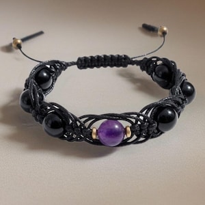 Puede incluir: Una pulsera de macramé negra con cuentas de ónix negro y una cuenta central de amatista púrpura. La pulsera tiene cordones negros ajustables con detalles dorados. El diseño es simétrico y las cuentas son lisas y pulidas.