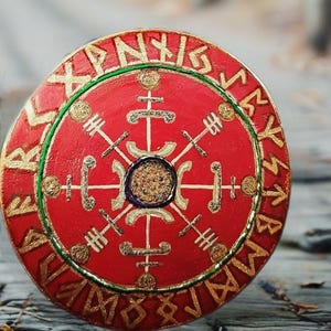 Puede incluir: Un escudo redondo rojo con símbolos rúnicos dorados y un diseño central. El escudo tiene un borde verde y está sobre un fondo de madera borroso. El diseño probablemente está inspirado en la cultura nórdica o vikinga.