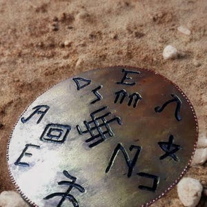 Może przedstawiać: Srebrzysty, owalny metalowy przedmiot z wytrawionymi symbolami i literami. Obiekt ma teksturowaną powierzchnię i miedzianą, koralikową krawędź. Spoczywa na piaszczystej powierzchni z małymi kamieniami.