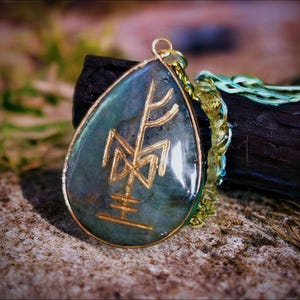 Könnte beinhalten: Tropfenförmiger Anhänger mit goldfarbenem Rahmen und goldfarbenem Runenmuster. Der Anhänger hat einen blau-grünen Stein in der Mitte und hängt an einer goldfarbenen Kette mit einem türkisfarbenen Band. Der Anhänger liegt auf einer rauen Oberfläche.