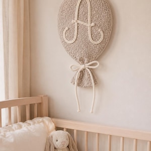 Peut inclure: Décoration murale en forme de ballon beige avec la lettre "A" blanche et un nœud noué. Le décor de la chambre d'enfant est fait d'un matériau doux et texturé et est suspendu au-dessus d'un lit bébé en bois avec un lapin en peluche.