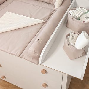 Könnte beinhalten: Eine beige Wickelauflage mit weißer, strukturierter Matte neben zwei beigen Stoffkörben auf einem weißen Regal. Die Körbe enthalten weiße gefaltete Tücher, eine Cremetube und eine Packung Feuchttücher mit sichtbarem Text auf der Verpackung.