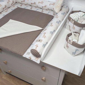 Könnte beinhalten: Ein weißer Wickeltisch mit einer Wickelauflage mit Giraffenmuster, einer braunen Waffelstoffmatte und zwei passenden Stoffkörben für Babyartikel. Die Körbe haben einen Giraffendruck und einen braunen Rand, passend zur Wickelauflage.