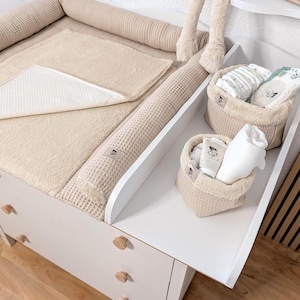 May include: A white changing table with a beige waffle-textured changing pad and two matching beige fabric storage baskets. The baskets contain baby care items including diapers, a white cloth, and a tube with a small label featuring a graphic.