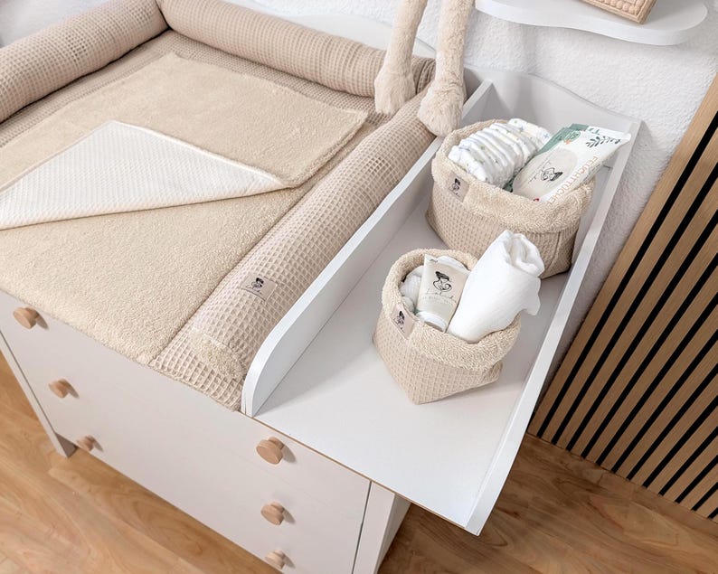 May include: A white changing table with a beige waffle-textured changing pad and two matching beige fabric storage baskets. The baskets contain baby care items including diapers, a white cloth, and a tube with a small label featuring a graphic.