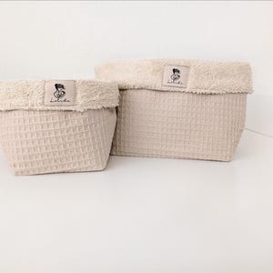 May include: Two beige storage baskets with a waffle-textured exterior and a soft, fuzzy, cream-colored folded rim. Each basket features a rectangular fabric label centered on the rim with a black line drawing of a person and the text Lolida.