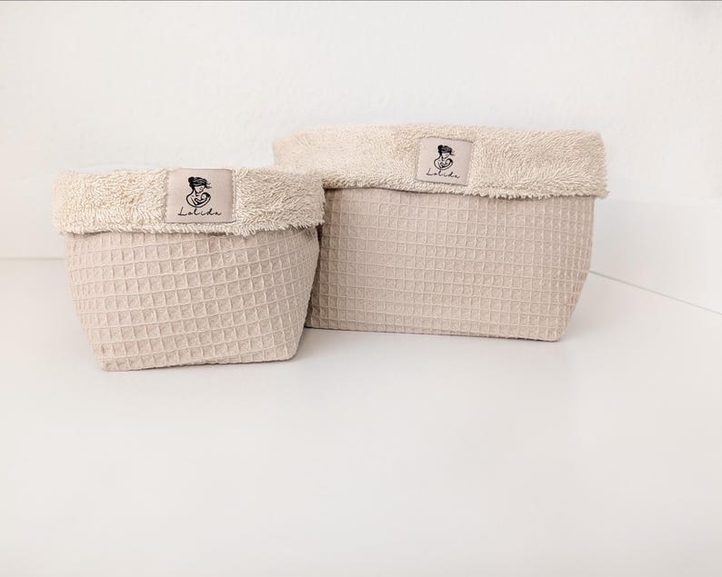 May include: Two beige storage baskets with a waffle-textured exterior and a soft, fuzzy, cream-colored folded rim. Each basket features a rectangular fabric label centered on the rim with a black line drawing of a person and the text Lolida.