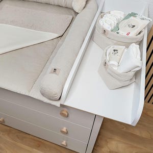 Può includere: Un fasciatoio bianco con imbottitura in velluto a coste beige chiaro e due cestini in tessuto abbinati. I cestini contengono panni bianchi, un tubetto di crema e una confezione di salviette. Il mobile ha tre cassetti con pomelli in legno.