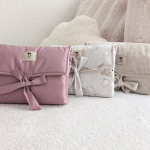 Op de afbeelding: Drie aankleedkussens in roze, wit met teddybeerprint en beige ribstof. Elk kussen heeft een striksluiting en een klein label. De kussens liggen op een wit, getextureerd oppervlak.