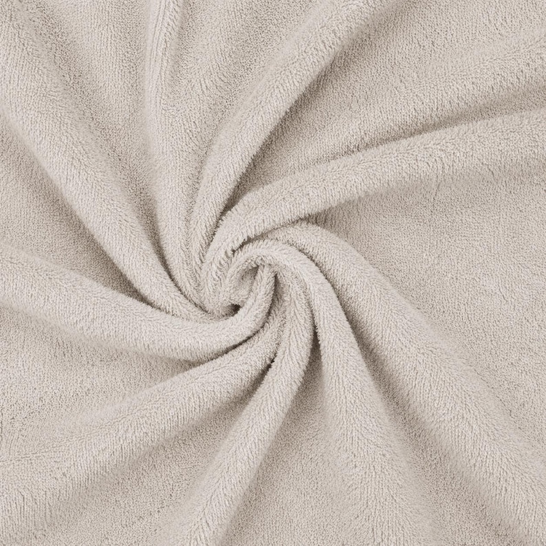 May include: A light beige towel featuring a dense, soft, and absorbent terry cloth texture. The fabric is arranged in a swirling, gathered pattern that highlights the looped pile construction and the consistent neutral color throughout the material.