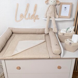 May include: A beige waffle-textured changing pad with raised side bolsters, featuring a matching beige towel overlay. Two beige fabric storage baskets with cream-colored trim sit on the white changing table next to a plush lamb toy.