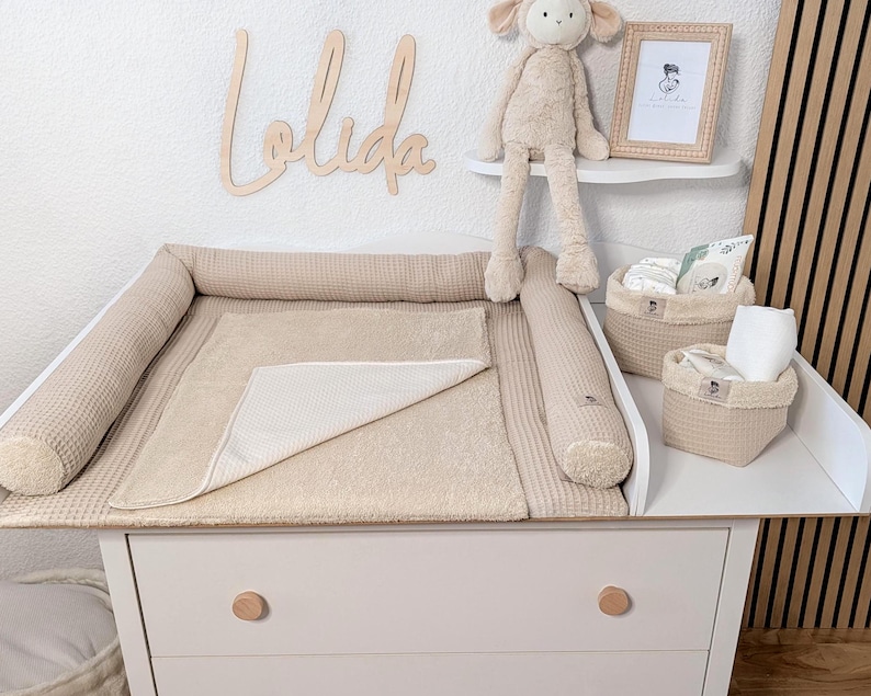 May include: A beige waffle-textured changing pad with raised side bolsters, featuring a matching beige towel overlay. Two beige fabric storage baskets with cream-colored trim sit on the white changing table next to a plush lamb toy.