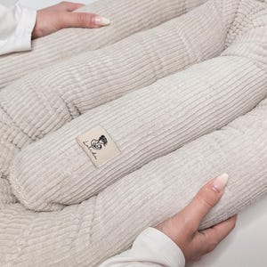 Könnte beinhalten: Ein beiges Babynest aus Kord mit weicher, gerippter Textur und einem U-förmigen, gepolsterten Rand. An der Seite ist ein rechteckiges Stoffetikett aufgenäht, das eine schwarze Strichzeichnung einer Person mit Baby und den Text Laluka zeigt.
