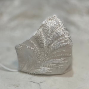 Puede incluir: Una mascarilla facial blanca con un delicado diseño de encaje con un patrón floral. El encaje está adornado con pequeñas cuentas blancas.