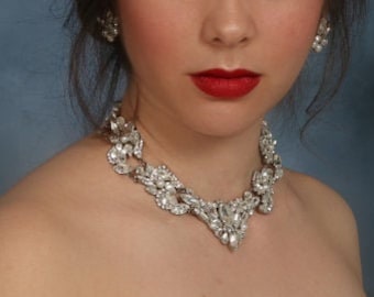 Op de afbeelding: Een vrouw met een sprankelende zilveren ketting en bijpassende oorbellen. De ketting is een opvallend sieraad met een gedetailleerd ontwerp. Ze draagt een strapless crèmekleurige jurk met kralenversieringen. Haar rode lippenstift zorgt voor een opvallend contrast.