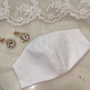 Puede incluir: Máscara facial de encaje blanco con un delicado patrón floral. La máscara está hecha de una tela suave y transpirable y tiene lazos elásticos para las orejas.