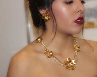 May include: A gold necklace with a double strand of chain and large flower-shaped charms. The necklace is worn by a person with dark hair and red lipstick.