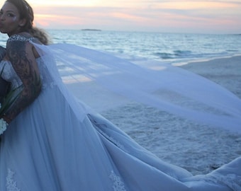 May include: A bride wearing a light blue wedding dress with a long veil blowing in the wind on a beach. The dress has lace detailing and the bride is holding a bouquet of white flowers.