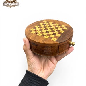 Op de afbeelding: Een ronde houten schaakset met een dambordpatroon op de bovenkant. De set is bruin met een goudkleurige metalen sluiting. Het logo "ANTIQUATEDERA" is zichtbaar. De set wordt in een hand gehouden tegen een witte achtergrond.