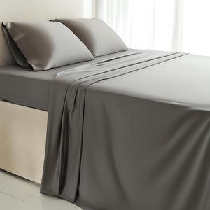May include: A bed with gray sheets and pillowcases. The bedding is neatly arranged, with the top sheet and pillowcases in place. The bed is against a neutral-colored headboard and a white curtain is in the background.