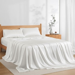 May include: A bed with a light wood frame is made up with a white sheet set. Two pillows are on the bed, and the sheet set drapes over the sides. A small wooden side table holds a vase with greenery.