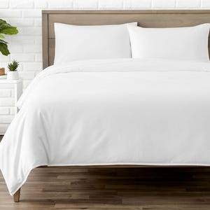 Può includere: Un set da letto bianco con un copripiumino e due federe. La biancheria da letto è disposta ordinatamente su un telaio del letto in legno. La stanza ha un'estetica minimalista con un muro di mattoni bianchi e pavimento in legno.