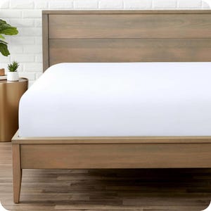 Könnte beinhalten: Ein Bett mit einem hellbraunen Holzrahmen und Kopfteil. Das Bett ist mit einem knackigen, weißen Spannbettlaken bezogen. Der Raum hat eine neutrale Farbpalette mit einer weißen Ziegelmauer im Hintergrund.