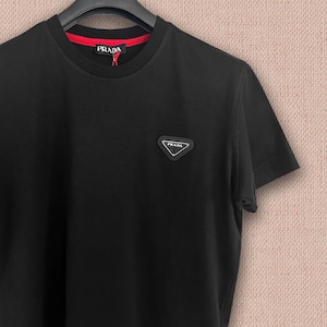 Könnte beinhalten: Ein schwarzes Kurzarm-T-Shirt mit Rundhalsausschnitt und rotem Innenkragen. Das Shirt zeigt einen schwarzen dreieckigen Aufnäher auf der Brust mit dem weißen Schriftzug Prada. Ein schwarzes Markenetikett mit weißem Schriftzug Prada ist am Hals.