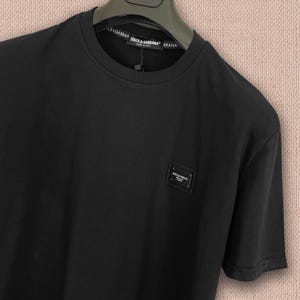 Puede incluir: Camiseta negra de cuello redondo con un pequeño parche cuadrado con el logo en el pecho. La camiseta de manga corta tiene un diseño sencillo y clásico. El nombre de la marca es visible en el cuello.