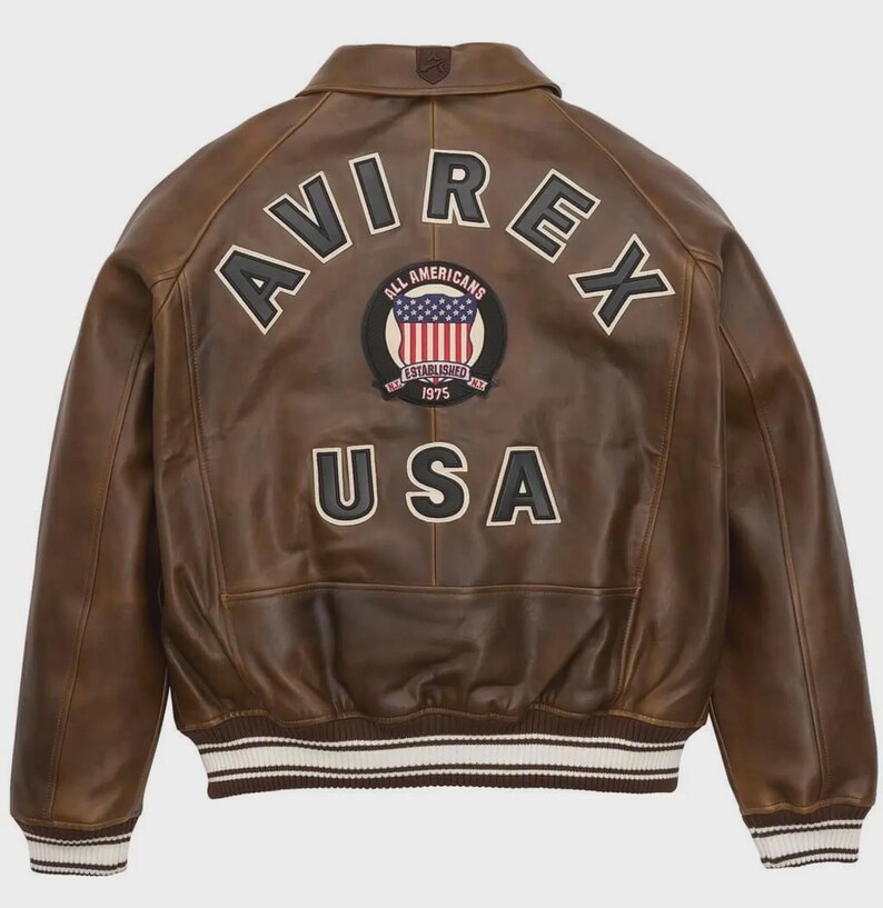 May include: Brown leather jacket with the word "AVIREX" arched across the shoulders in white block letters. Below is a circular patch with the American flag and the words "ALL AMERICANS ESTABLISHED 1975". The word "USA" is below the patch.
