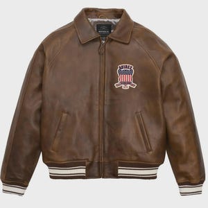 May include: Brown leather jacket with a zip-up front, collared neckline, and ribbed cuffs and hem. Features a patch with the text "AVIREX" and an American flag design. The jacket has two front pockets.