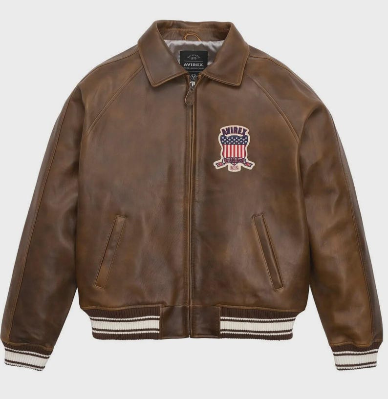 May include: Brown leather jacket with a zip-up front, collared neckline, and ribbed cuffs and hem. Features a patch with the text "AVIREX" and an American flag design. The jacket has two front pockets.