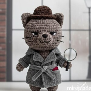May include: A crocheted cat detective doll wearing a gray trench coat, brown hat, and holding a magnifying glass. The cat has black whiskers and a red pocket square. The doll is standing in front of a window.
