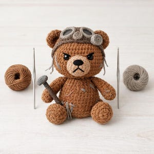 Puede incluir: Un oso de ganchillo marrón hecho a mano con un sombrero de aviador marrón y un clavo gris. Dos ganchillos y ovillos de lana en marrón y gris están a ambos lados del oso. El oso tiene una expresión enfadada.