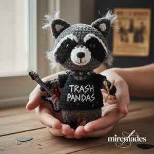 May include: Crocheted raccoon doll in gray, black, and white, wearing a black t-shirt with "Trash Pandas" text and a studded collar. The raccoon holds a miniature guitar and drumsticks, a unique handmade item.