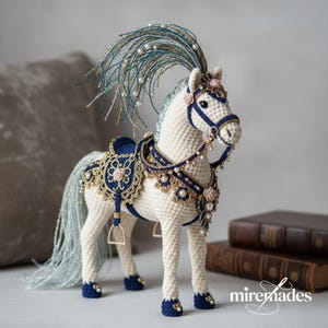 Może przedstawiać: Ręcznie wykonana figurka konia z szydełka w kolorze kremowym z misternymi granatowymi i złotymi zdobieniami. Koń posiada ozdobne siodło, uzdę oraz płynącą grzywę i ogon. Uzupełniony perłami i małymi różowymi różami, ma około 25 cm wysokości.