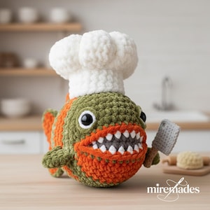 May include: A crocheted piranha wearing a chef's hat and holding a small cleaver. The fish is green and orange with a wide, toothy grin. The chef's hat is white. The background is a kitchen setting.