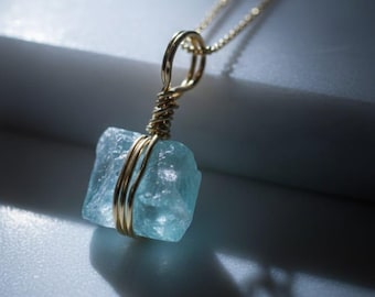 May include: A gold-colored chain necklace with a pendant featuring a light blue, raw gemstone. The gemstone is wrapped in gold-colored wire. The necklace is displayed on a light gray surface.